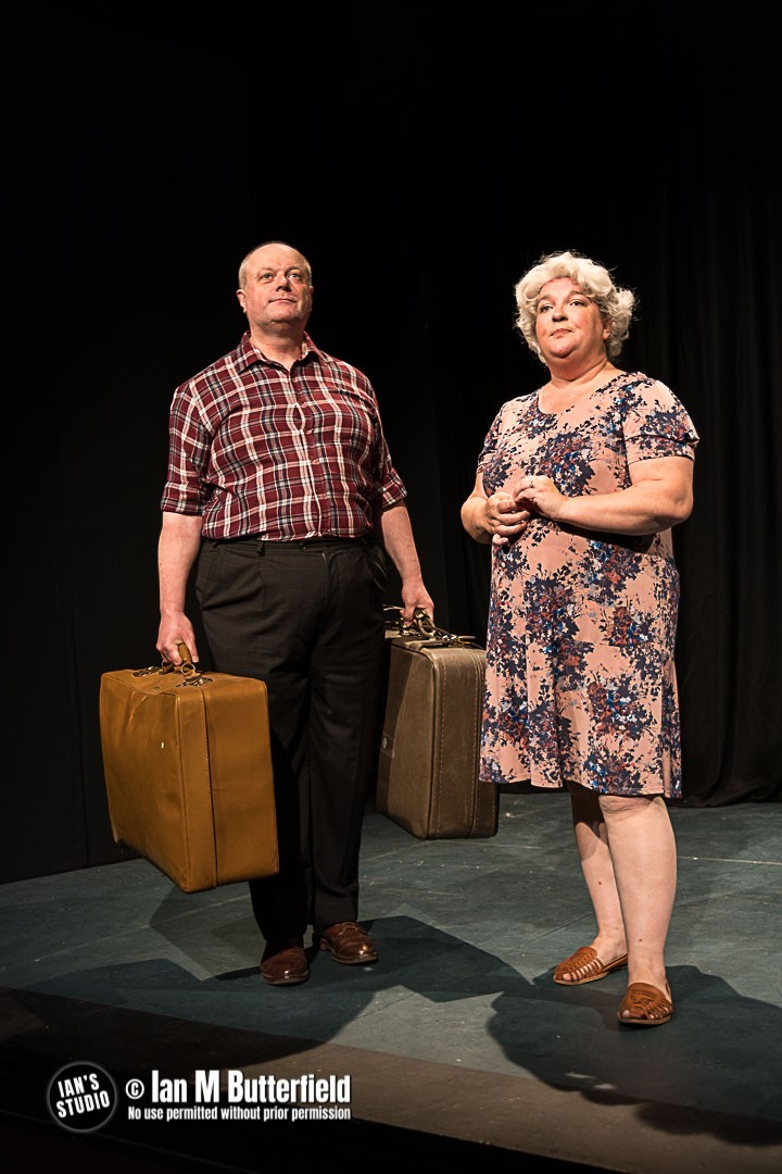A man and women stood on a black stage with an old fashioned suitcase