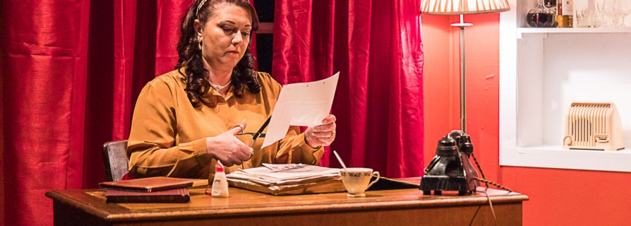 A stage. A women sat at a desk cutting some paper. There is an old fashioned phone on the desk.
